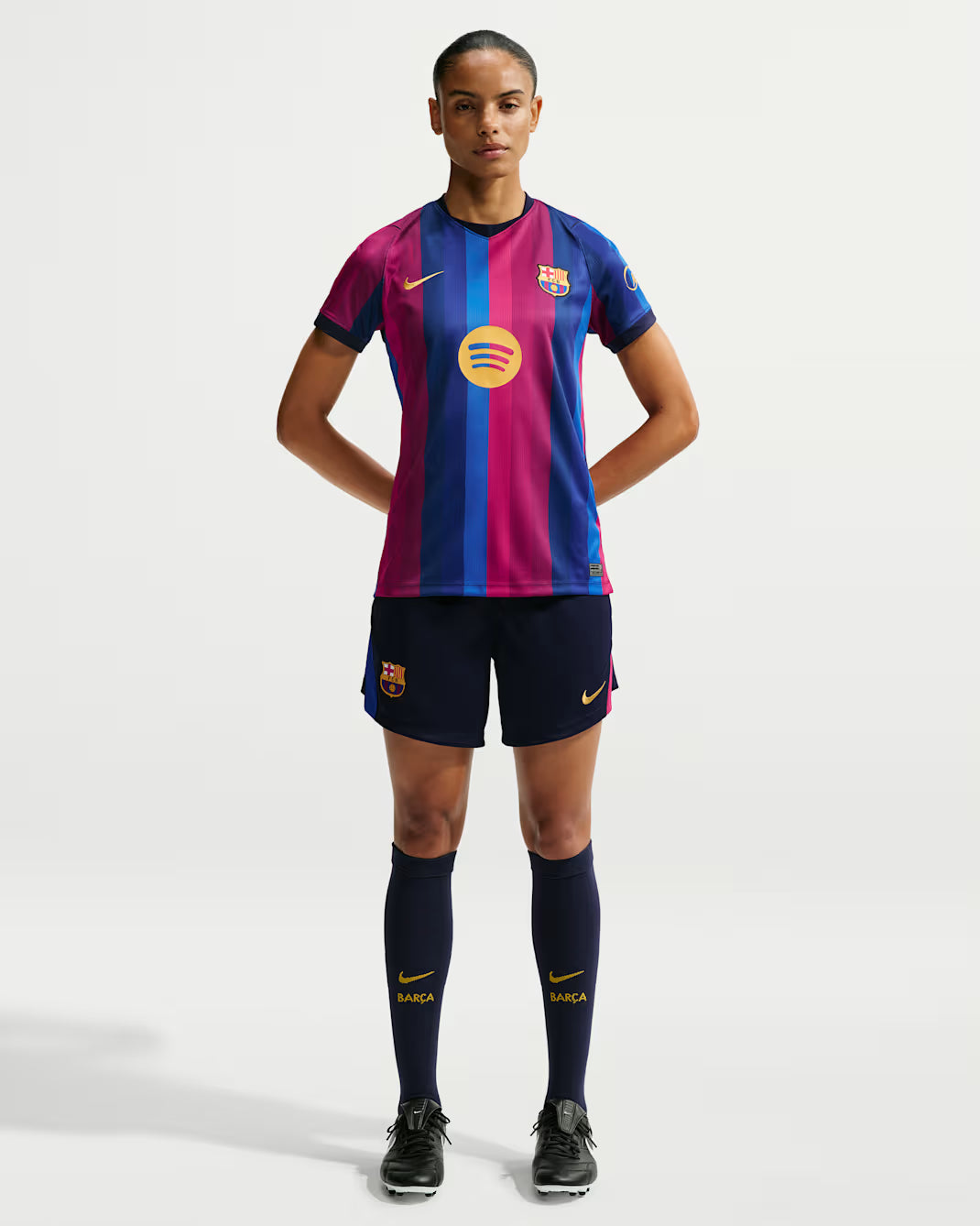 A female model stands against a light gray studio background wearing an FC Barcelona home soccer jersey, navy shorts, navy socks, and black cleats. The jersey has vertical magenta and blue stripes with a yellow circular logo in the center and a small yellow Nike swoosh on the chest.