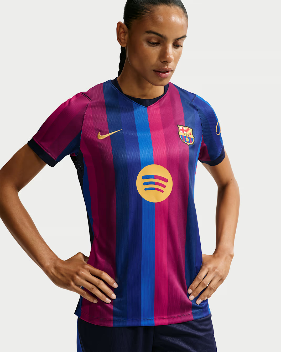 A woman wearing a Nike FC Barcelona home soccer jersey with vertical magenta and blue stripes, featuring the yellow Spotify sponsor logo on the chest, set against a light gray studio background.