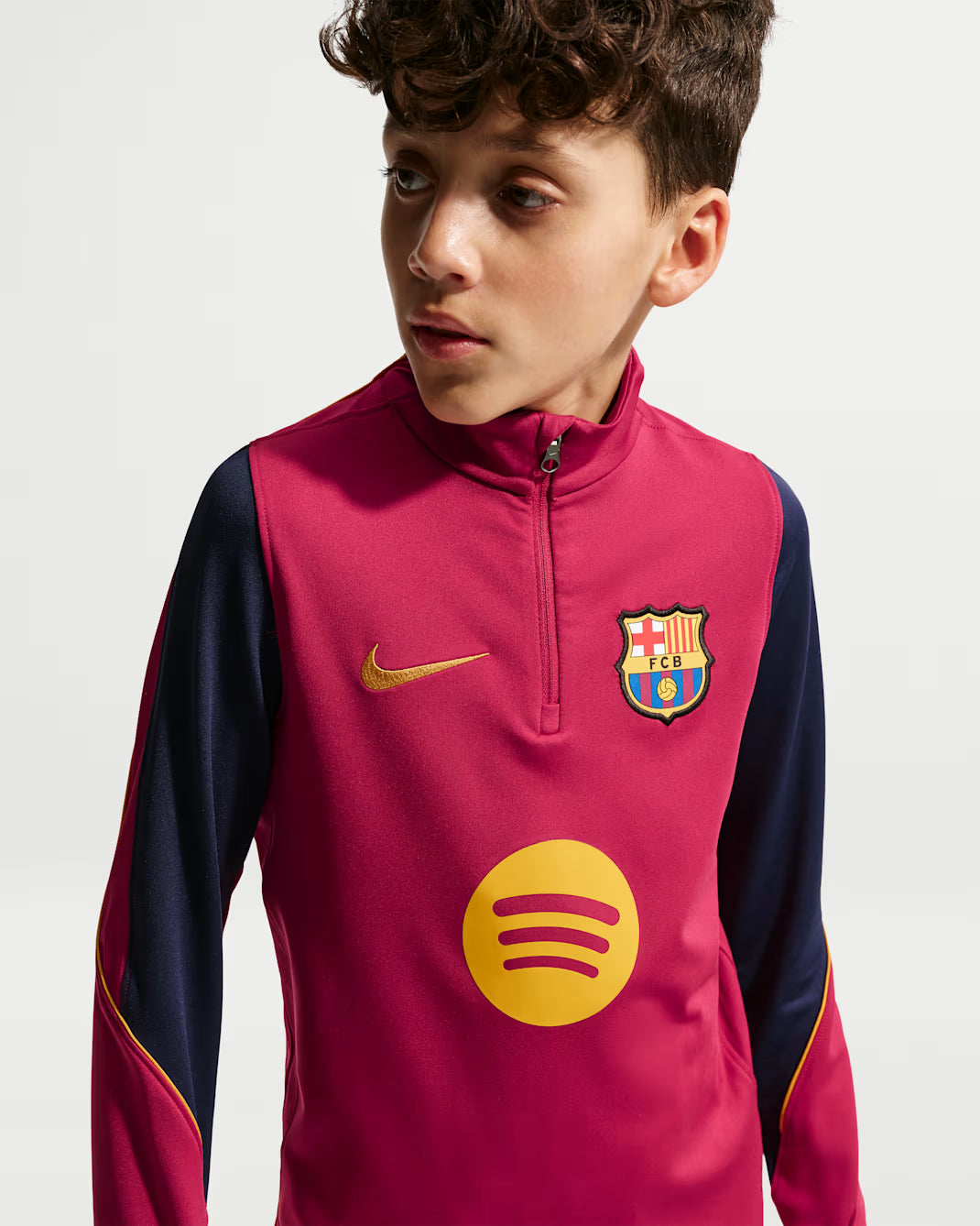 A young boy wearing a pink and navy blue FC Barcelona training top with a yellow Nike swoosh and a yellow Spotify sponsor logo, photographed against a light gray studio background.