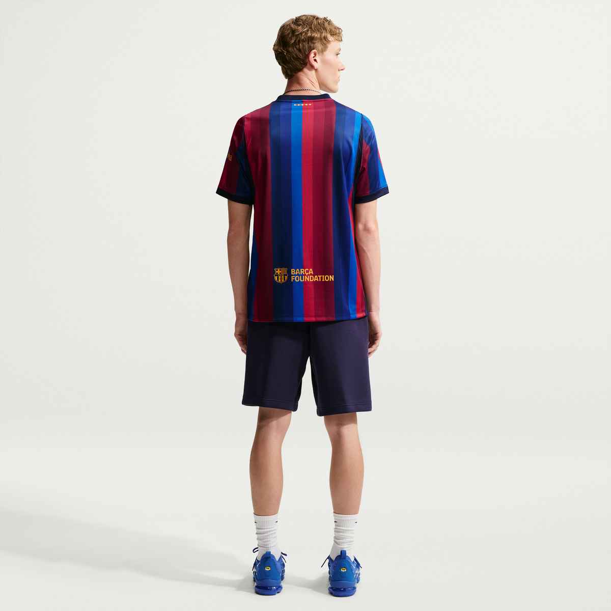 A young male model shown from the back wearing a striped FC Barcelona soccer jersey, navy shorts, white socks, and blue cleats against a light gray studio background.