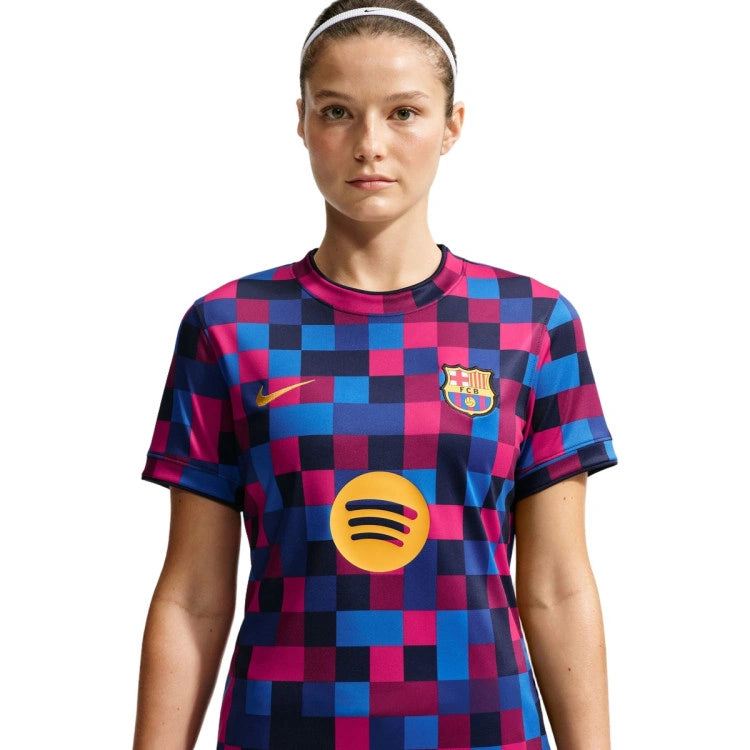 A woman modeling a short-sleeve soccer jersey with a pink, blue, and navy pixelated check pattern. The jersey features a yellow Nike swoosh on the right chest, the FC Barcelona crest on the left chest, and a large yellow Spotify logo on the center.