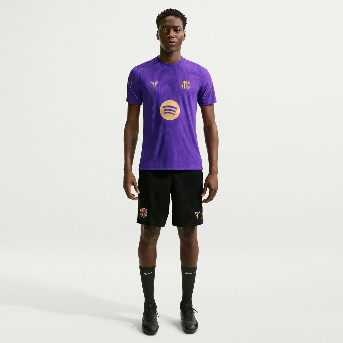 A male model stands against a light gray studio background wearing a purple short-sleeve soccer jersey with gold logos, black shorts, black socks, and black cleats. The jersey has a large circular gold logo on the front and a smaller crest on the chest.