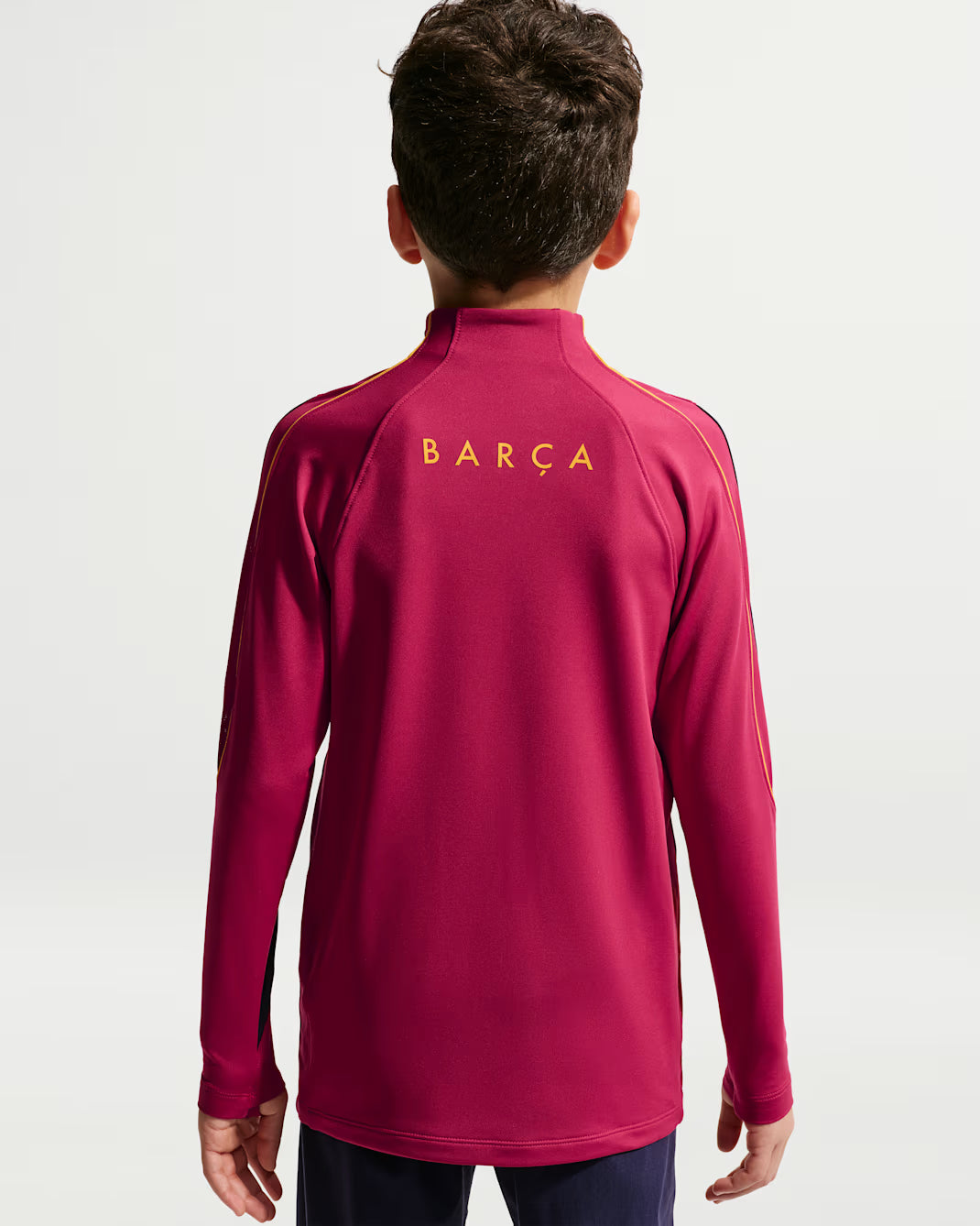 A child viewed from behind wearing a magenta long-sleeve athletic top with 'BARÇA' printed in gold on the upper back, against a light gray studio background.