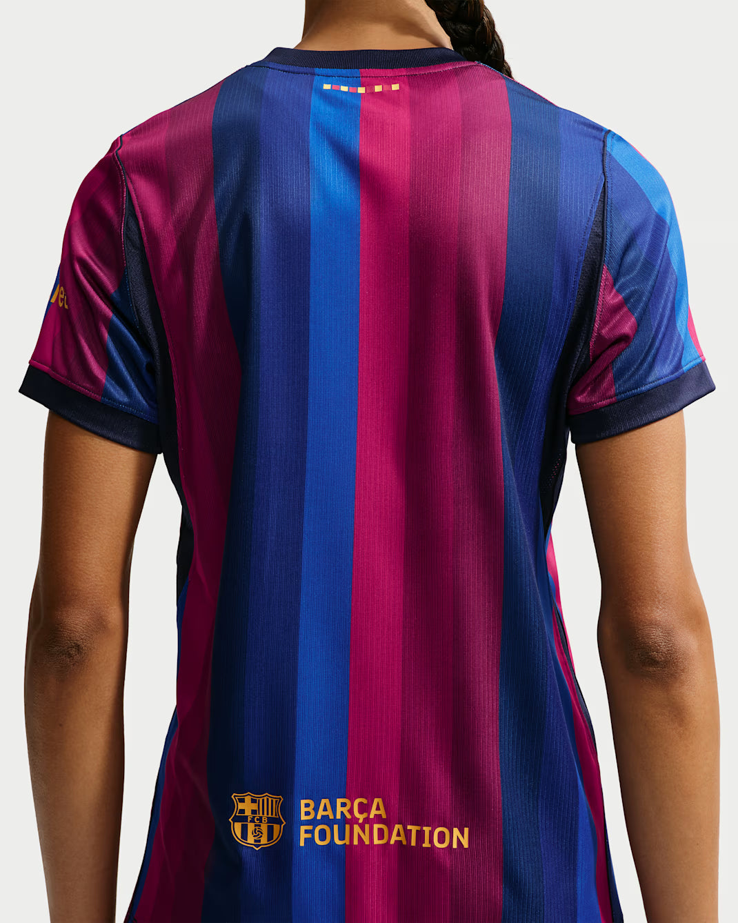 Back view of a short-sleeve FC Barcelona jersey with vertical magenta and blue stripes, navy trim, and a gold FC Barcelona Foundation logo near the lower back.
