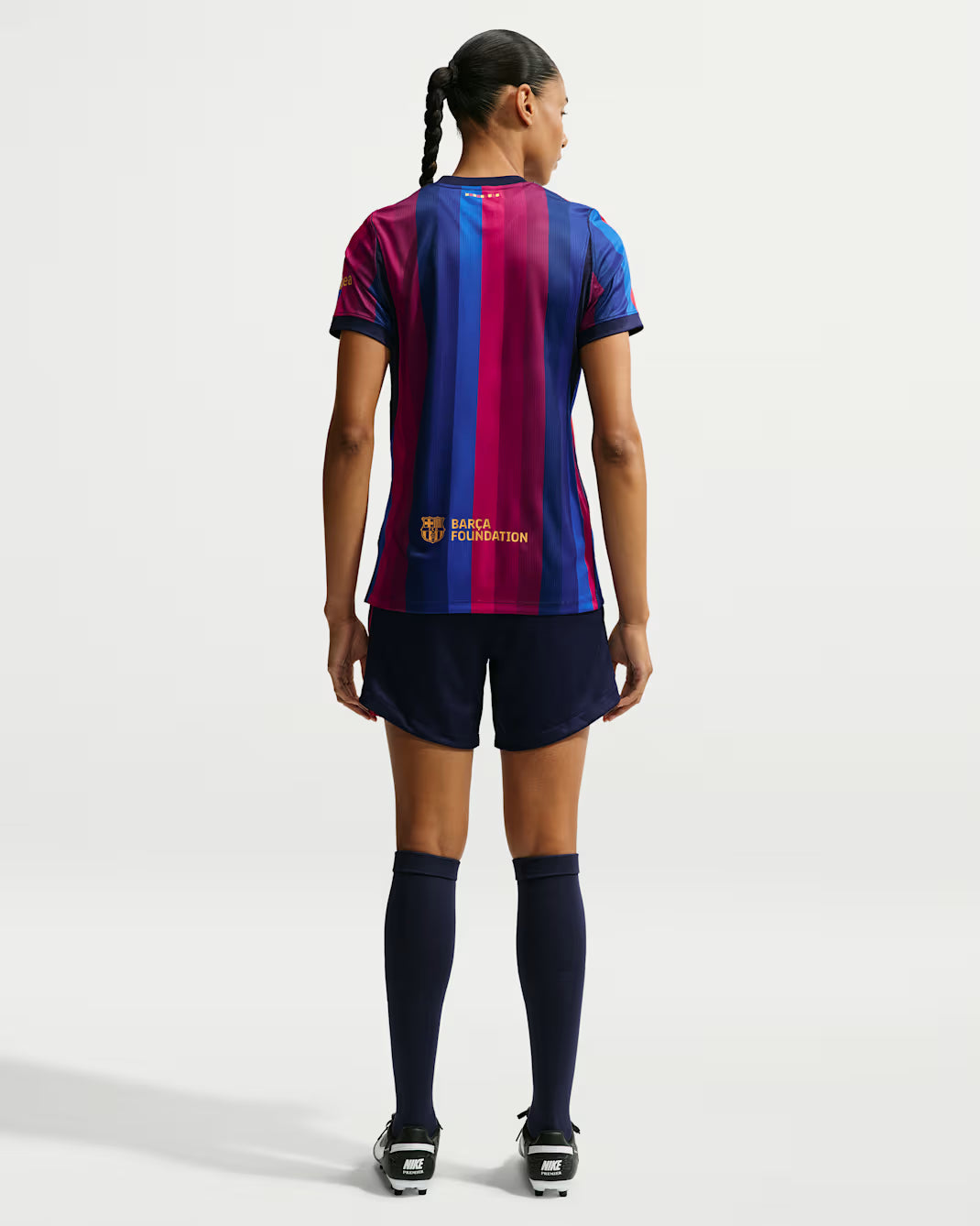 A model shown from the back wearing a short-sleeve soccer jersey with vertical blue and magenta stripes, navy shorts, navy knee-high socks, and black cleats against a light gray studio background.