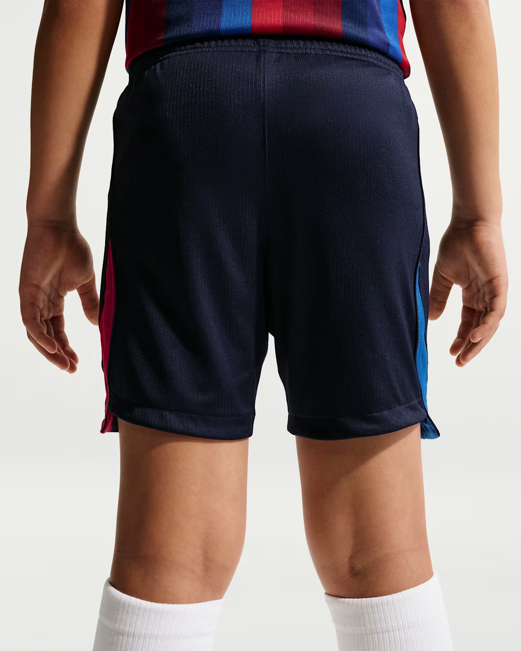 Rear view of a child wearing dark navy athletic shorts with pink and blue side stripes, paired with a striped jersey and white socks, against a light gray studio background.