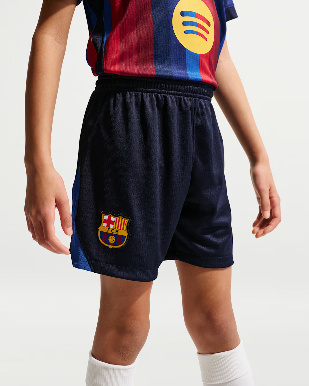 A child wearing dark navy FC Barcelona home soccer shorts with the club crest on the left leg, paired with a matching striped jersey and white socks, shown against a light gray studio background.