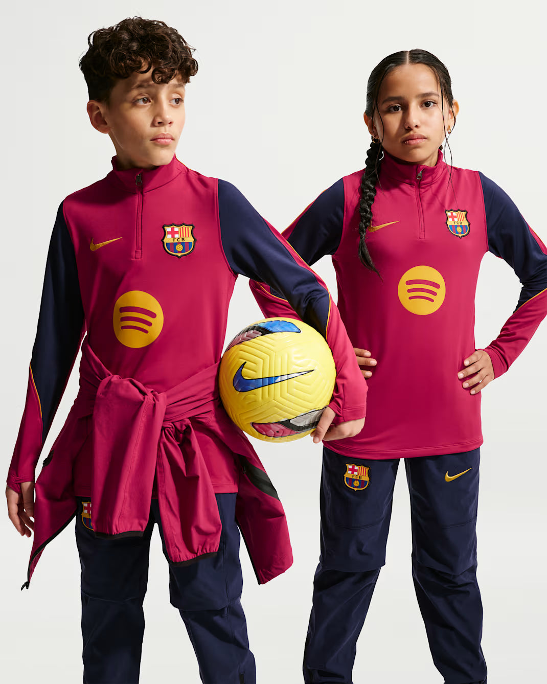 Two children modeling matching FC Barcelona athletic tracksuits with a pink and navy colorway, featuring the club crest, Nike swoosh, and yellow Spotify sponsor logo. The boy holds a yellow Nike soccer ball against a clean white studio background.